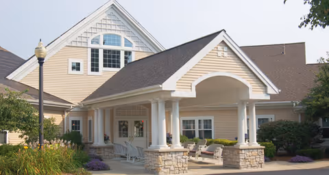Exterior view of a beige senior living facility building with a covered entrance supported by white columns and stone bases. There are rocking chairs and potted plants on the porch, with landscaping including bushes and flowers around the building.