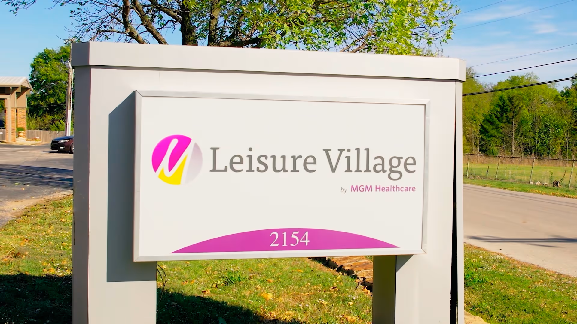 A white outdoor sign for Leisure Village (2154) with a pink and yellow logo standing by a road and grassy area.