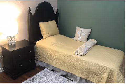 A small bedroom with a single bed covered in a yellow quilt and a pillow with a decorative design. A rolled towel is placed on the bed. Next to the bed is a dark wooden nightstand with two drawers and a lit table lamp with a white shade. The walls are painted white and green, and there is a light-colored rug on the wooden floor.