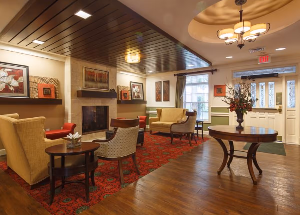 Warmly furnished senior living lobby with seating around a fireplace, a round table with flowers, and an entrance door.
