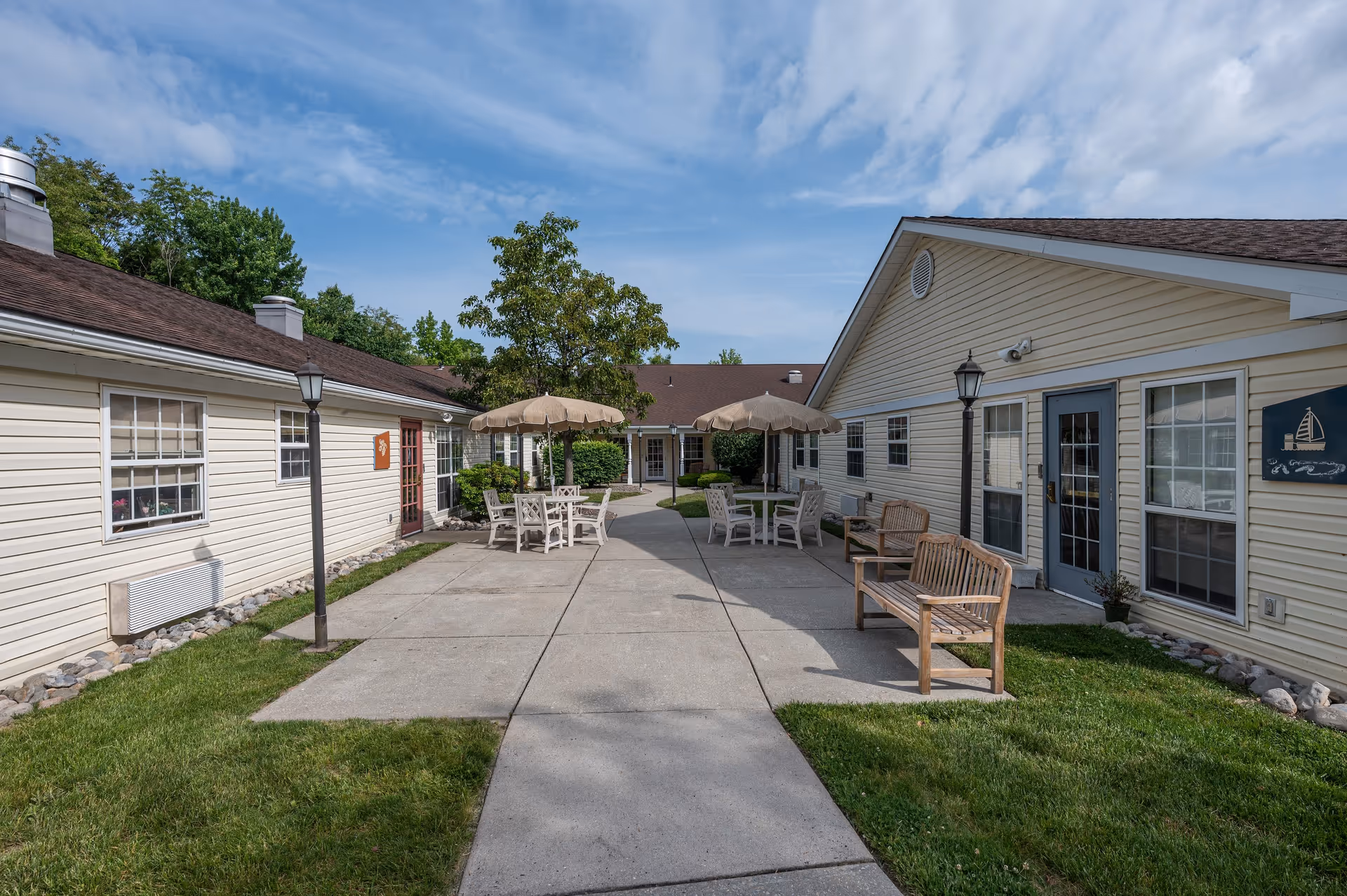 Outdoor courtyard area of a senior living facility with beige siding buildings on both sides, concrete walkways, green grass, wooden benches, and tables with umbrellas. Trees and a partly cloudy sky are visible in the background.