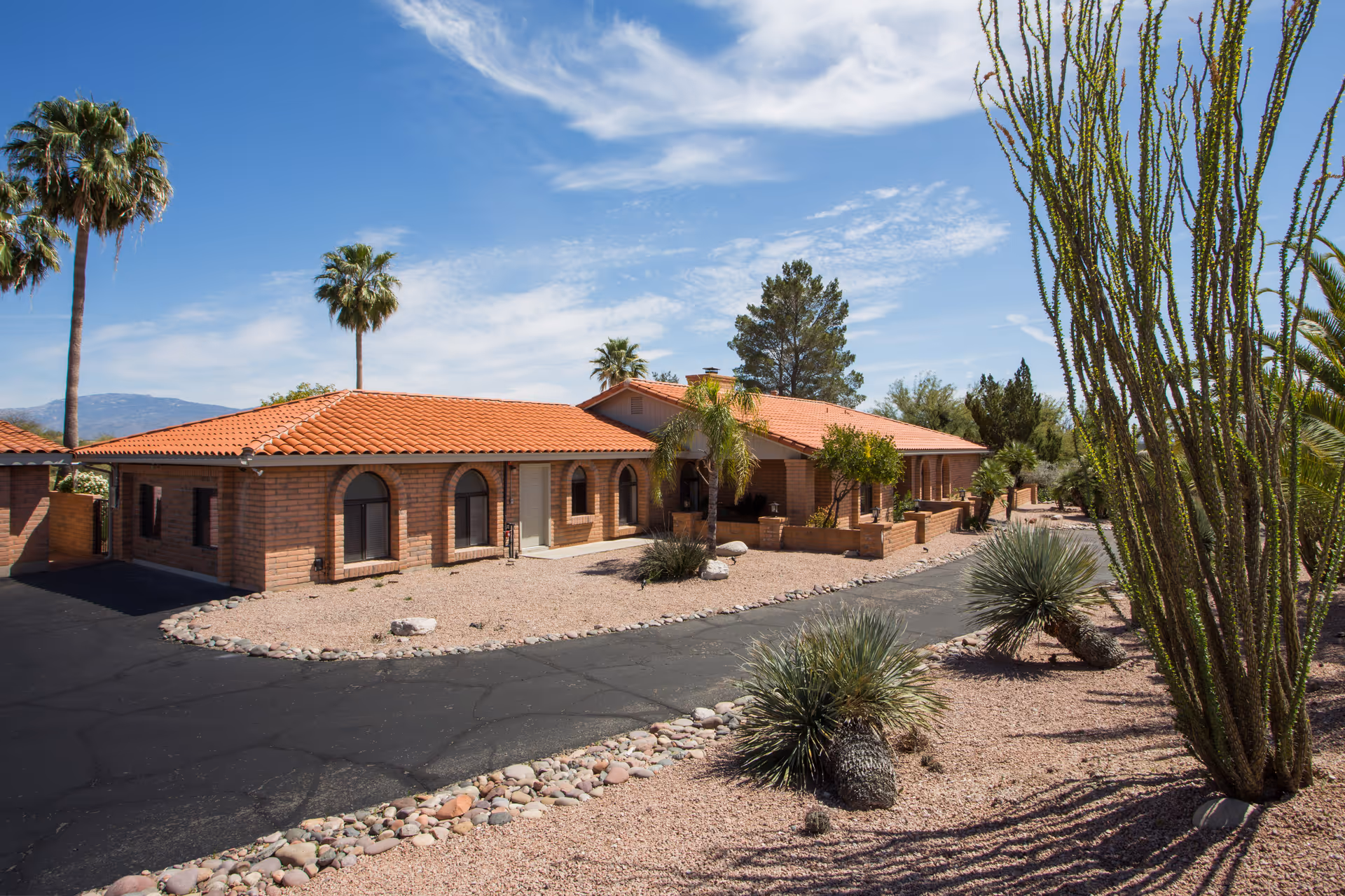 Exterior view of a single-story building with a red tile roof and brick walls, surrounded by desert landscaping including palm trees, cacti, and gravel. The sky is clear with some clouds.
