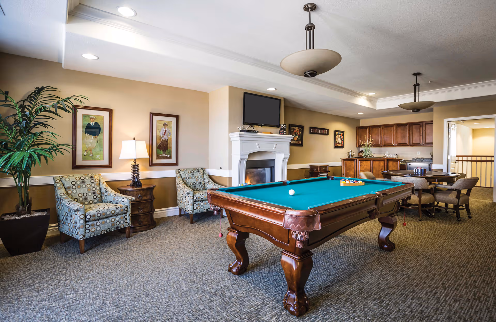 A senior living facility common room featuring a pool table with balls set up for a game, two patterned armchairs with a small wooden side table and lamp between them, a large potted plant, a fireplace with a TV mounted above it, and a kitchenette area with cabinets and a round table with chairs.