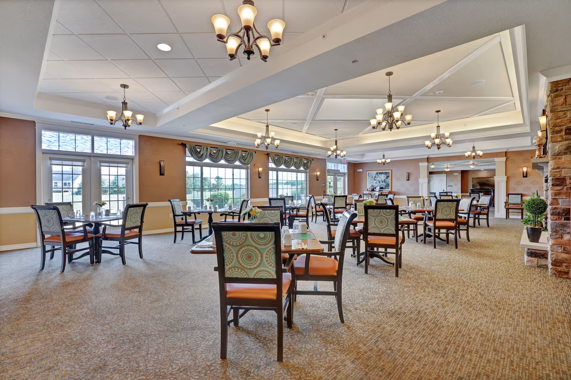 Spacious dining room with multiple tables and chairs arranged neatly. The room features large windows allowing natural light to fill the space, elegant chandeliers hanging from a coffered ceiling, and a stone fireplace on the right side. The walls are painted in warm tones, and the carpeted floor adds to the cozy atmosphere.