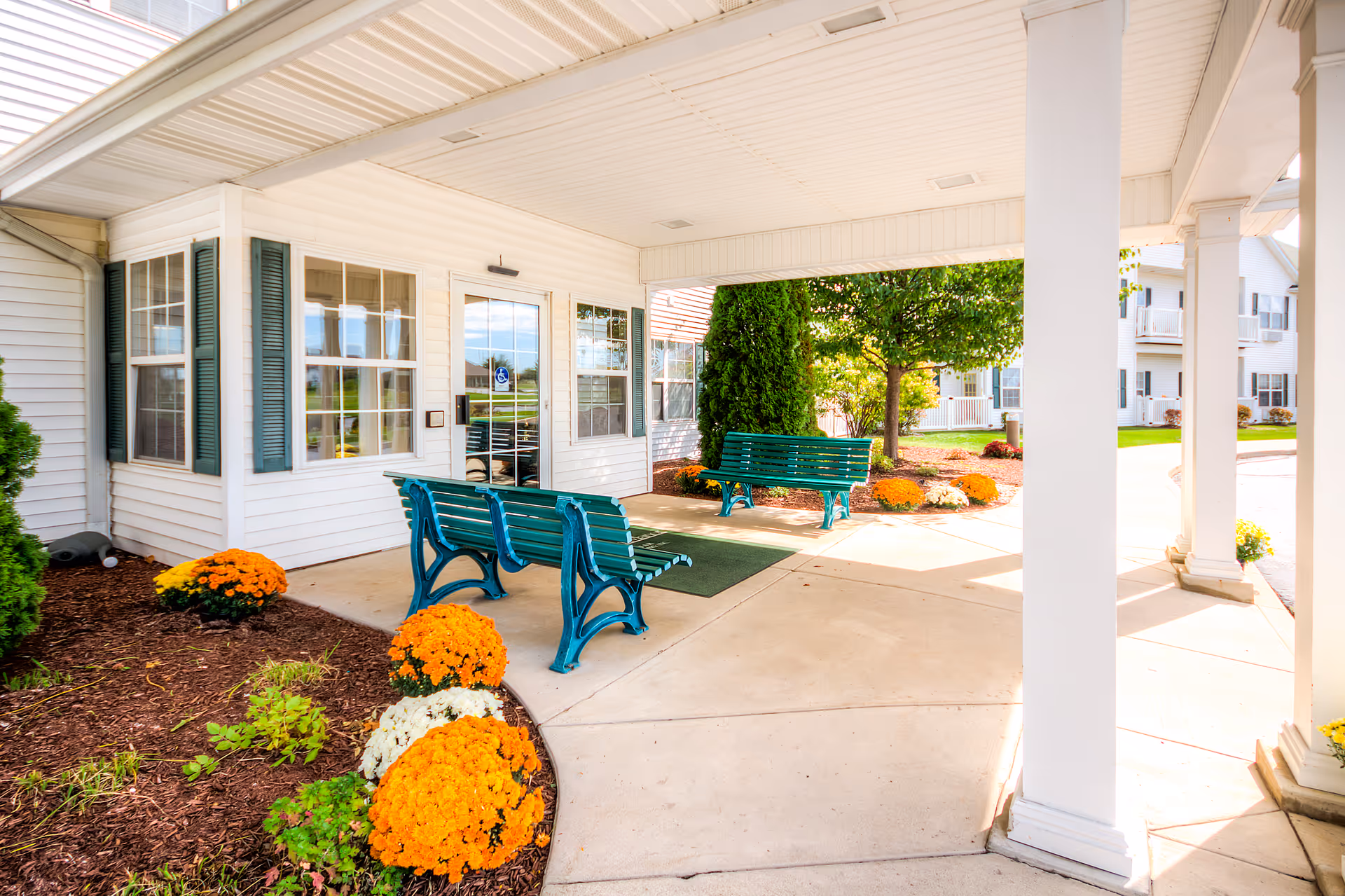 Covered outdoor seating area with green benches and a concrete floor at the entrance of a white building with green shutters. There are orange and white flowers planted in mulch beds around the area, and a tree and other greenery are visible in the background.