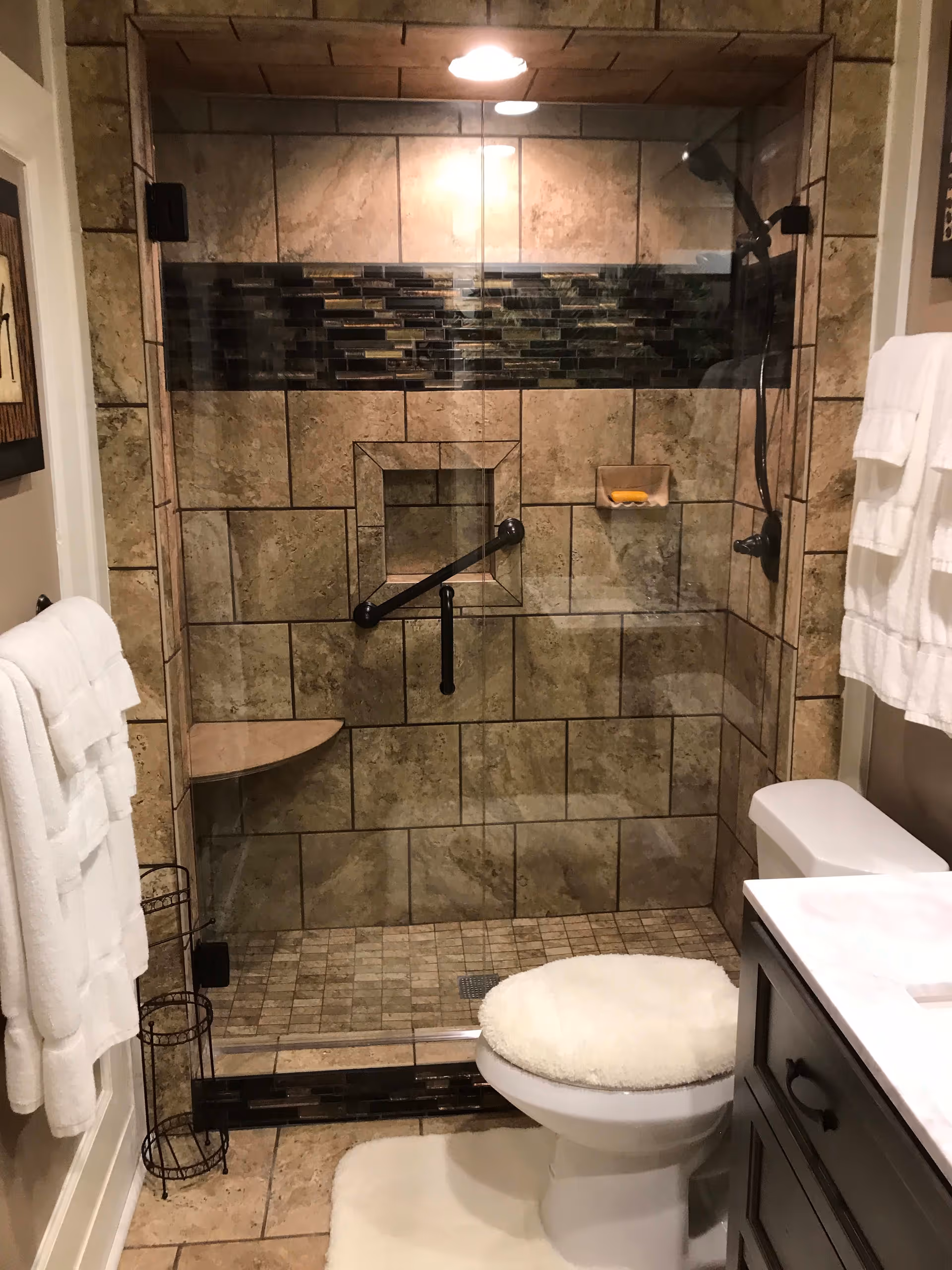 A bathroom with a walk-in shower featuring beige and brown tiled walls and floor, a glass door, a built-in corner seat, and a grab bar. There is a toilet with a fluffy white seat cover and matching white rug in front. White towels hang on racks on the walls, and a dark wood vanity with a white countertop is partially visible.