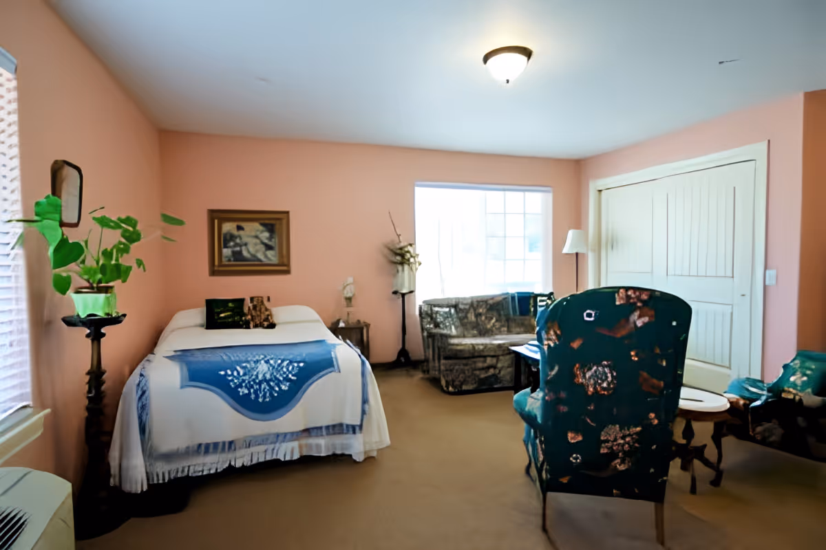 A cozy bedroom with peach-colored walls featuring a bed with a white and blue blanket, a framed picture above the bed, a plant on a tall stand near the window, a small side table with a lamp, a floral-patterned armchair, a loveseat, a floor lamp, and a closet with double doors.