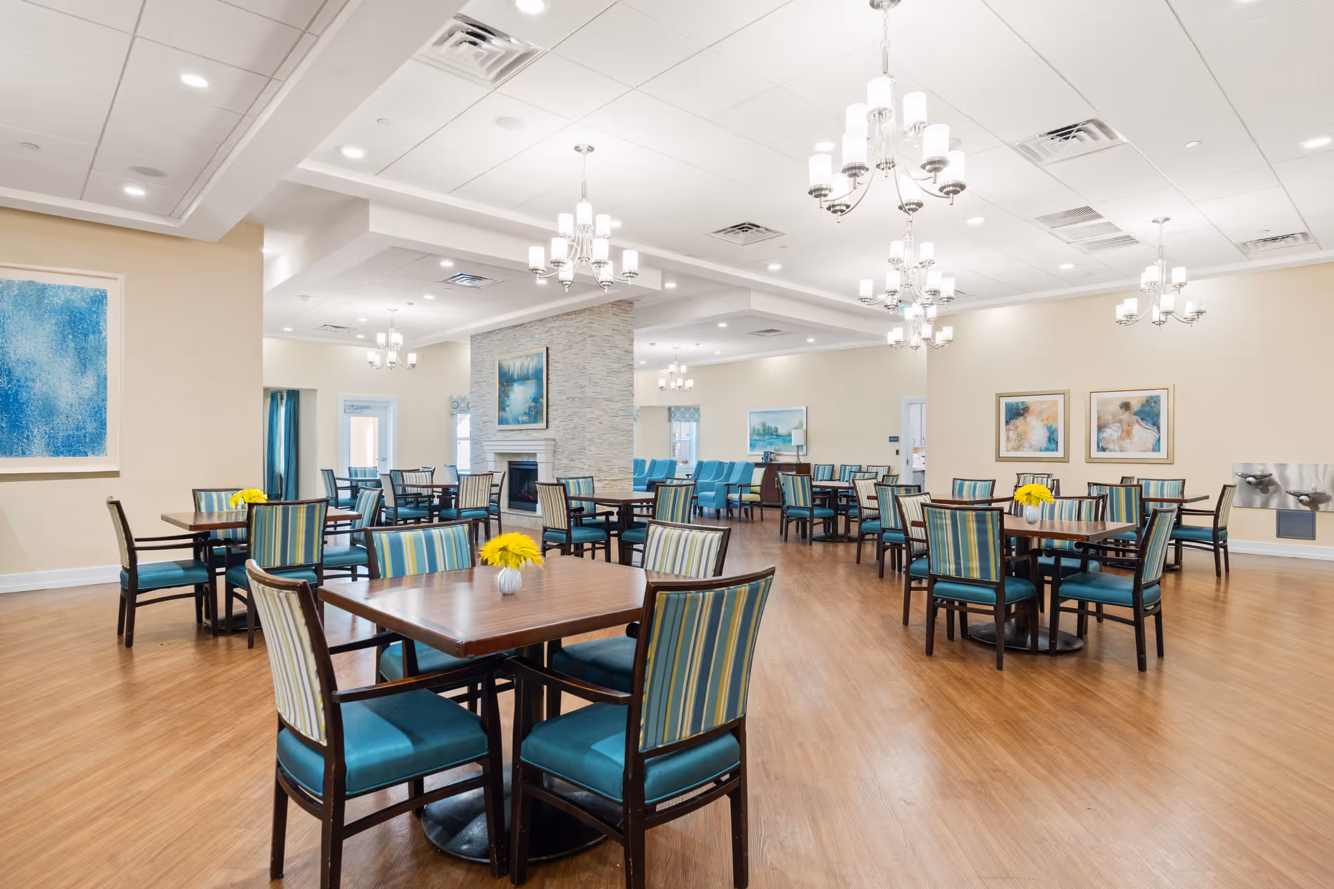A spacious dining room with multiple square wooden tables surrounded by teal and striped upholstered chairs. The room features a light wood floor, beige walls adorned with blue-themed artwork, and several elegant chandeliers hanging from a white ceiling. A stone fireplace is visible in the center of the room, and there are water fountains mounted on one wall.
