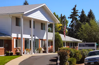 Exterior view of Laramie Care Center building with white siding and brick accents, surrounded by trees and bushes. An American flag is visible near the entrance, and a sign with the facility's name is placed near the driveway where a car is parked.