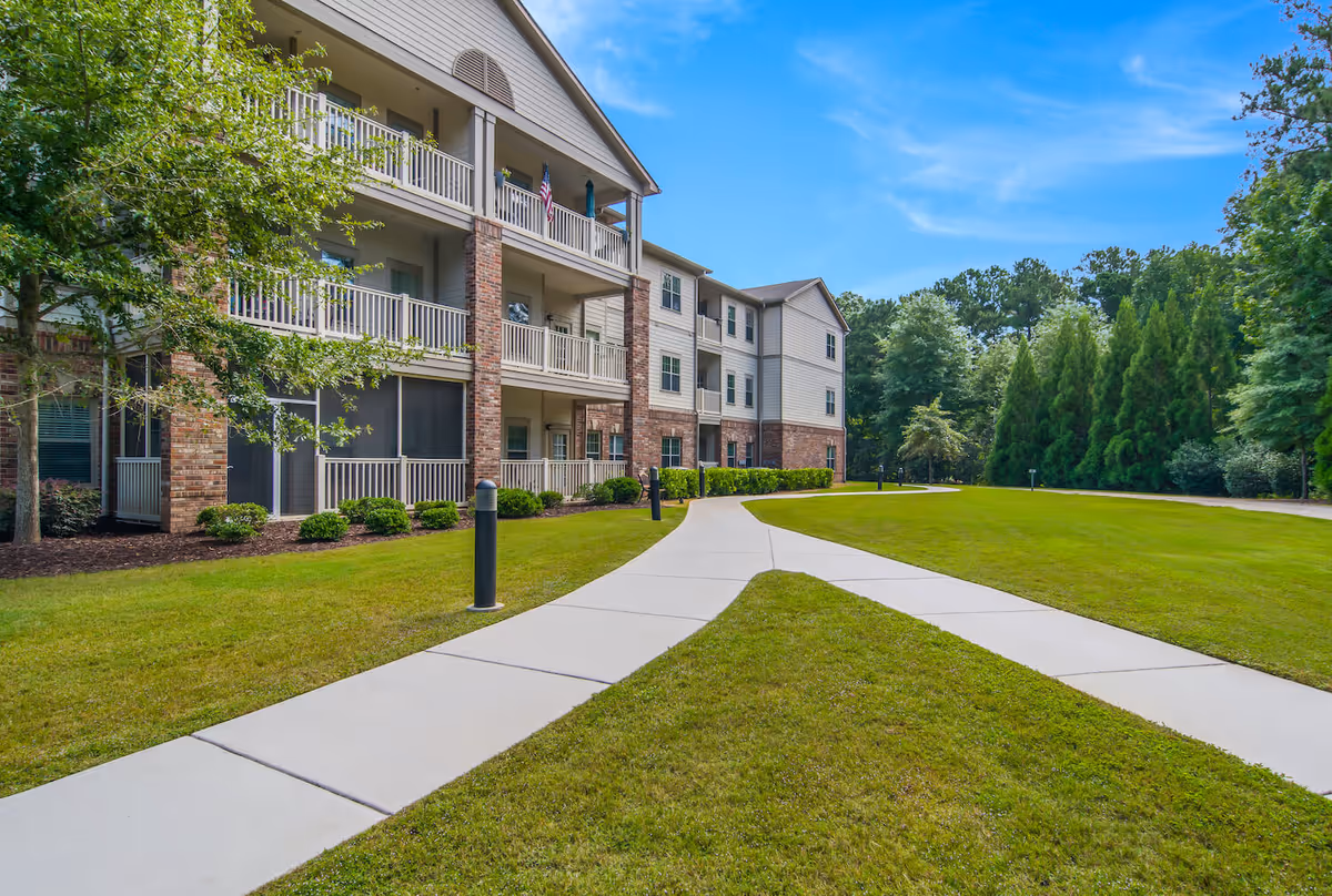 Three-story senior living building with balconies next to a paved walkway and manicured lawn under a blue sky.