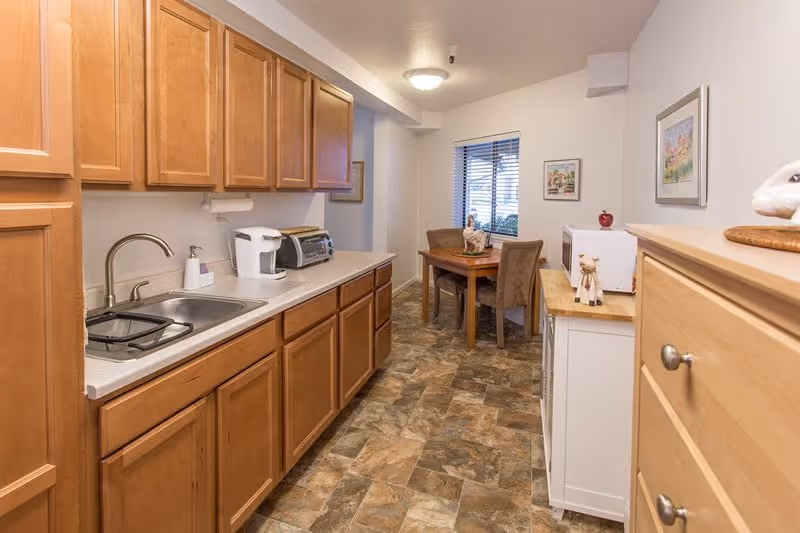 A narrow kitchen area with wooden cabinets on the left side, a countertop with a sink, a coffee maker, and a toaster oven. At the far end, there is a small dining table with two chairs near a window with blinds. On the right side, there is a white cabinet with a microwave on top and a wooden dresser. The floor has a patterned tile design, and the walls are light-colored with framed artwork.