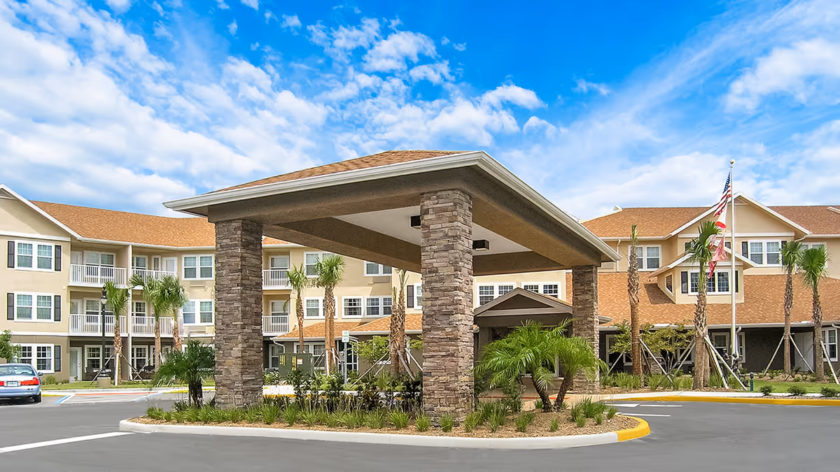 Exterior view of a senior living facility with a covered entrance supported by stone pillars, surrounded by palm trees and landscaping, under a partly cloudy blue sky.