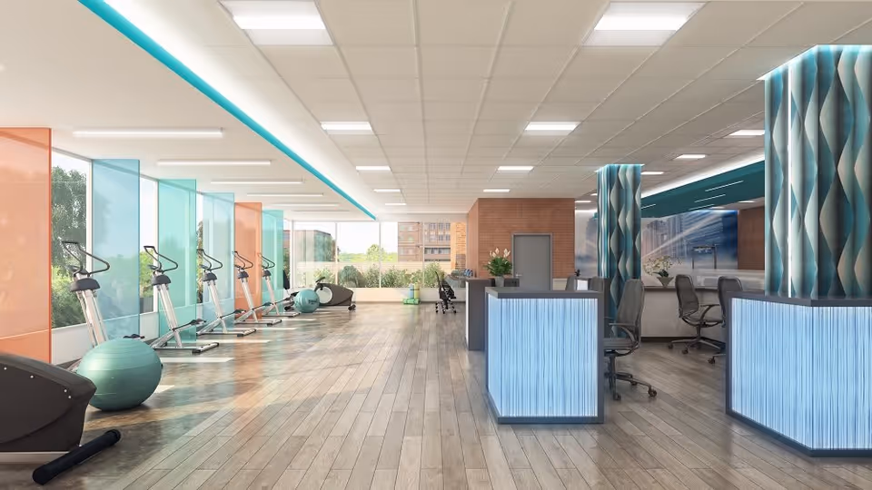 A spacious, modern fitness center with exercise equipment including elliptical machines and stability balls along the left side near large windows. The room has wood flooring, bright overhead lighting, and a reception desk with illuminated panels on the right side. Decorative columns with a blue and white wave pattern are also visible.