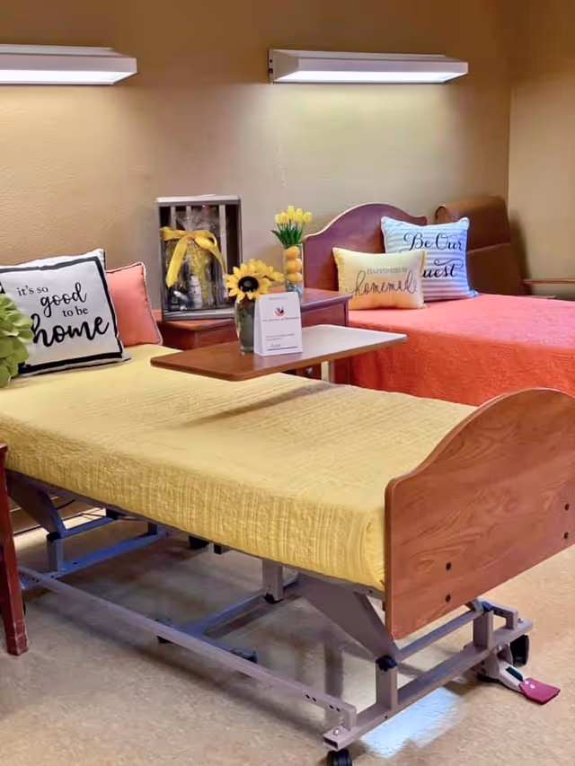 A cozy room with two beds, one with a yellow bedspread and the other with an orange bedspread. Each bed has decorative pillows with welcoming messages like 'it's so good to be home', 'Be Our Guest', and 'happiness is homemade'. There is a small table between the beds with a vase of yellow flowers and a card. The room has warm lighting and a comfortable atmosphere.