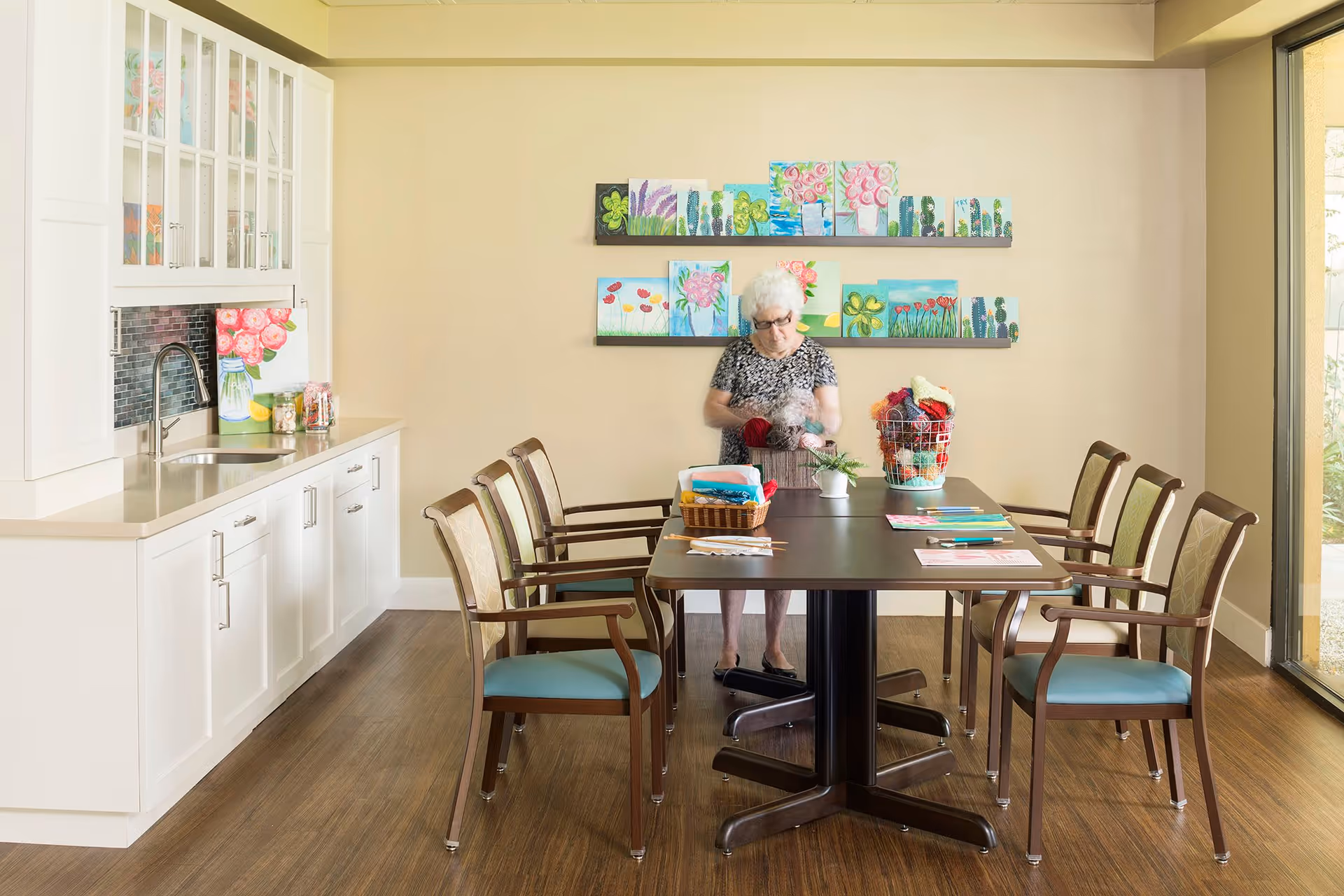 A senior woman standing at a rectangular table in a well-lit room with wooden flooring. The table is surrounded by six chairs with wooden frames and light blue cushions. On the table are various craft supplies and a basket filled with yarn. Behind the woman, colorful floral paintings are displayed on the beige wall. To the left, there is a white cabinet with a sink and glass-front upper cabinets. Large windows on the right side let in natural light.