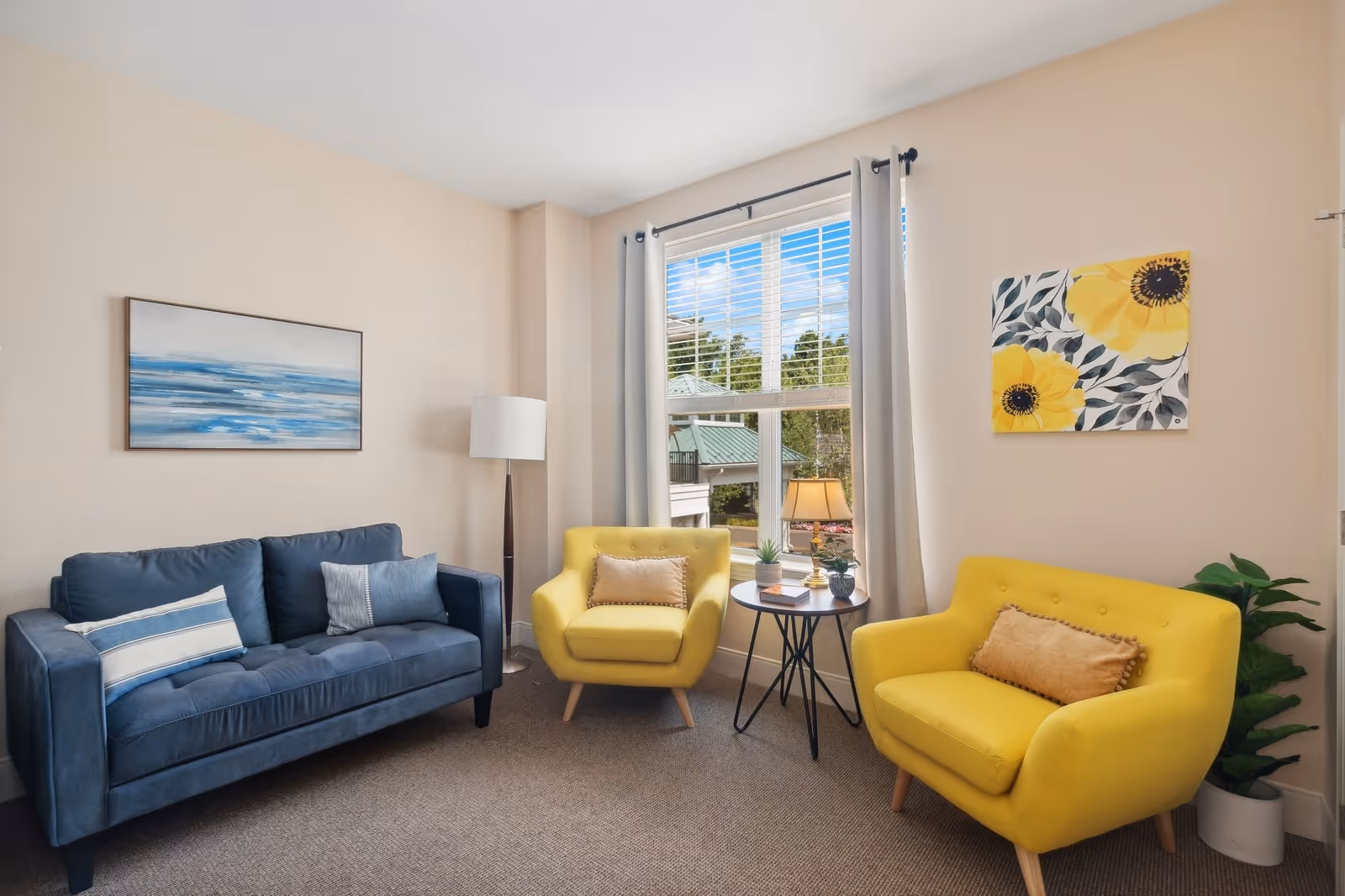 A cozy living room with a blue sofa and two yellow armchairs arranged around a small round table. The room has beige walls with two paintings, one depicting blue waves and the other yellow flowers. A large window with white blinds lets in natural light, and there is a floor lamp and a potted plant in the corners.