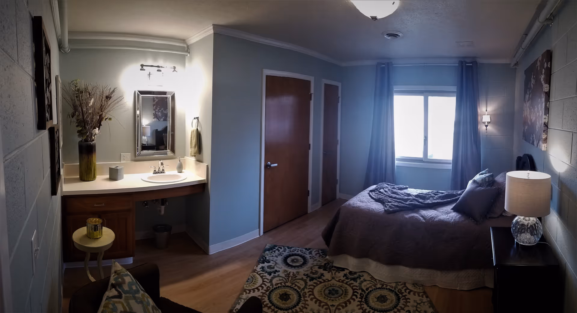 A cozy bedroom in a senior living facility featuring a bed with pillows and a blanket, a bedside table with a lamp, a window with curtains, two closed wooden doors, a small vanity with a sink, mirror, and decorative vase with dried plants, and a patterned area rug on the wooden floor.