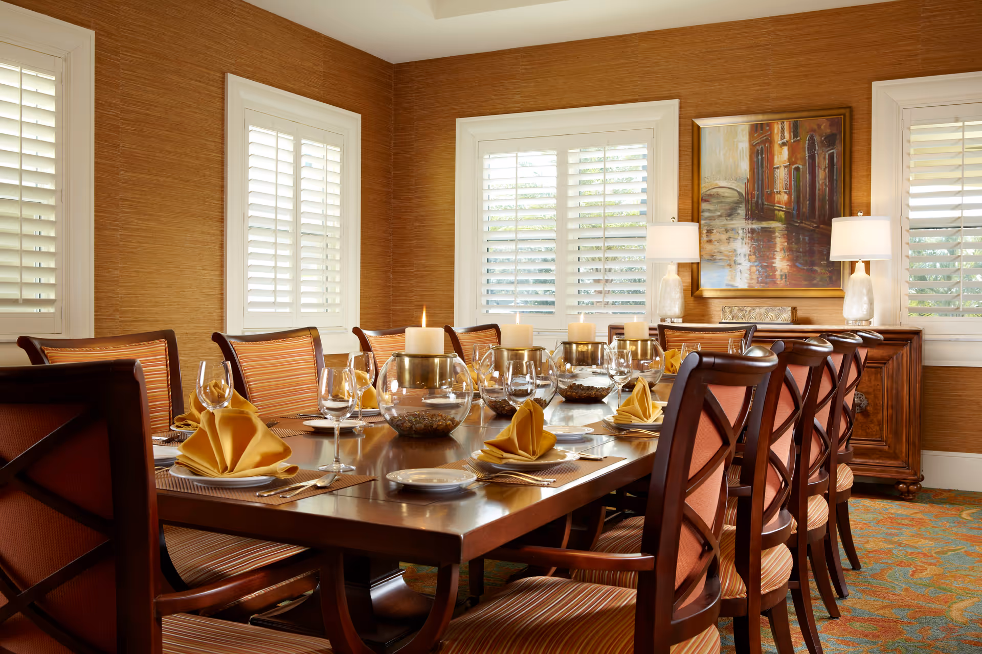 Formal dining room with a long wooden table set with folded yellow napkins, glassware, and candles surrounded by upholstered chairs in a warm, well-lit interior.