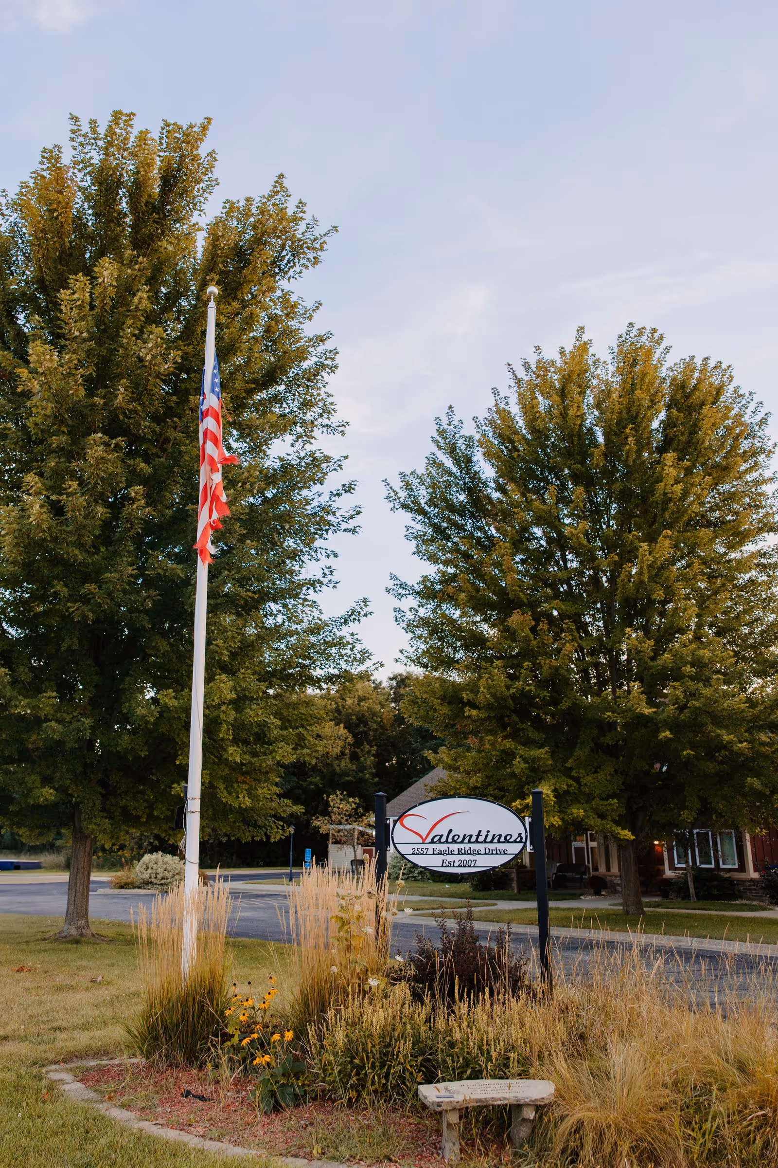 Outdoor view of Valentines Assisted Living facility sign surrounded by tall grasses and flowers, with an American flag on a flagpole and large trees in the background under a partly cloudy sky.