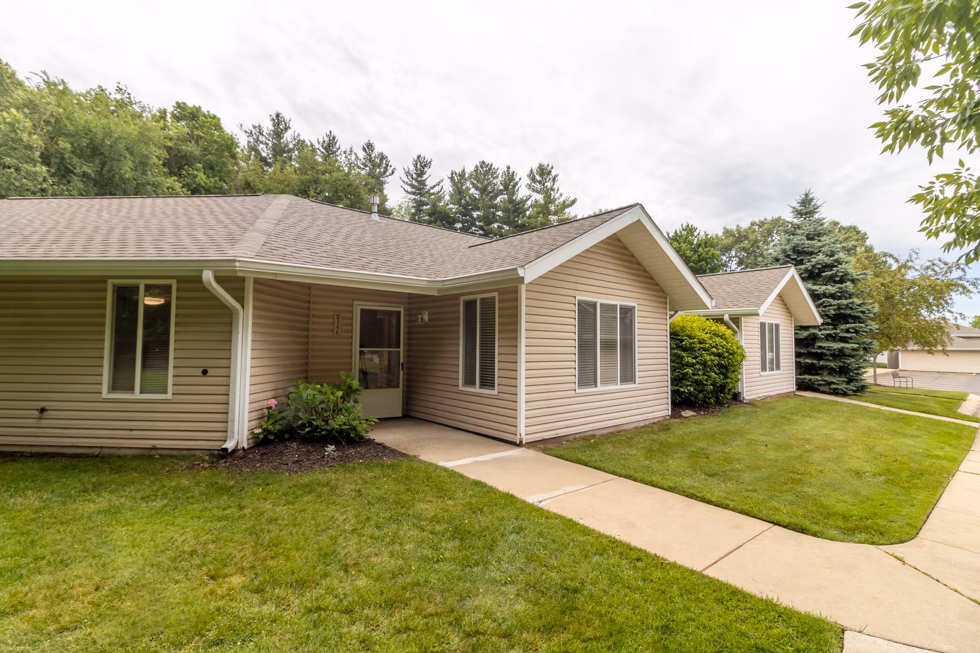 Exterior view of a single-story residential building with beige siding, white trim, and a shingled roof. The building is surrounded by a well-maintained lawn, bushes, and trees. A concrete walkway leads to a door under a small covered porch area. The sky is overcast.