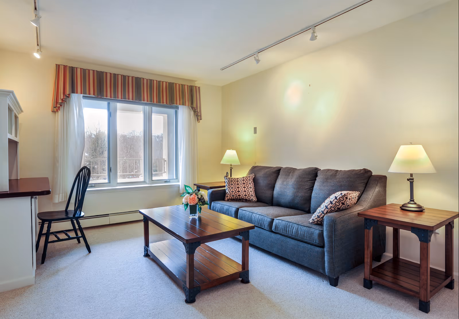 A cozy living room with a gray sofa adorned with two patterned pillows, a wooden coffee table with a small flower arrangement, two wooden side tables each with a lamp, a black wooden chair, and a large window with striped valance and white curtains letting in natural light.