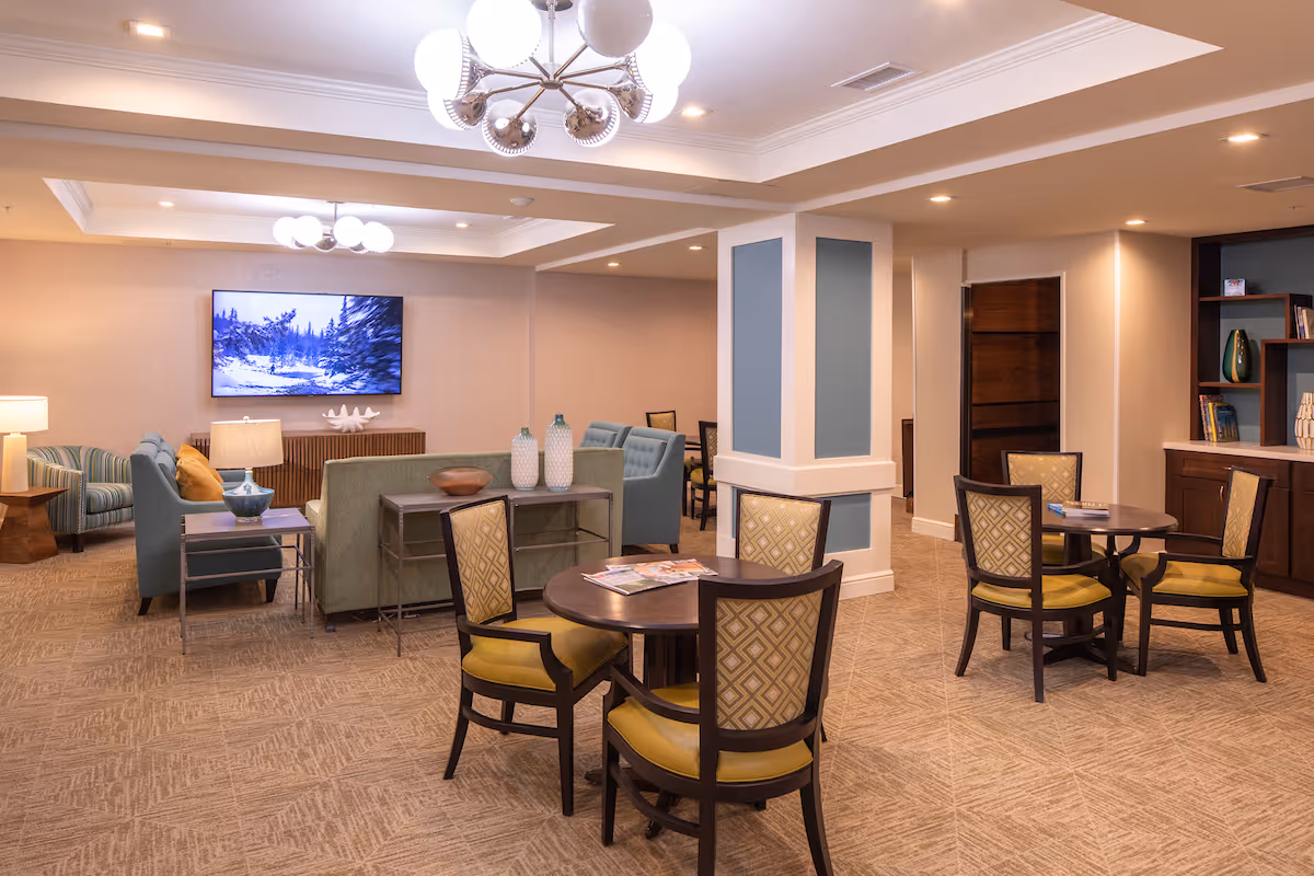 A spacious and well-lit common area in a senior living facility featuring multiple seating arrangements including round tables with chairs and sofas facing a wall-mounted television. The room has beige walls, carpeted floors, modern light fixtures, and decorative vases on tables and shelves.
