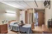 A clean patient room with an adjustable hospital bed, an overbed table holding a laptop, wooden bedside furniture, a privacy curtain, and a wall-mounted TV.