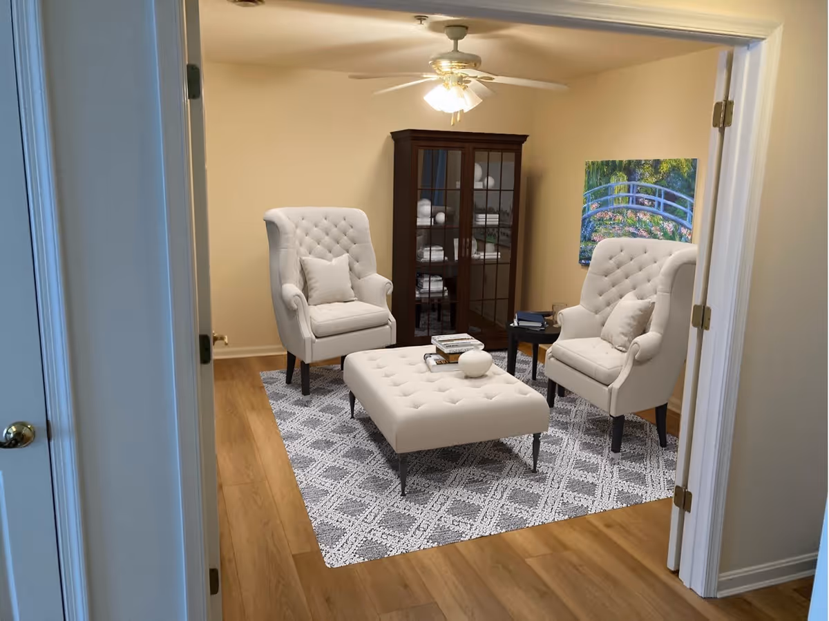 View through an open doorway into a living room with two tufted armchairs, a tufted ottoman on a patterned rug, a wooden cabinet, wall art and a ceiling fan.