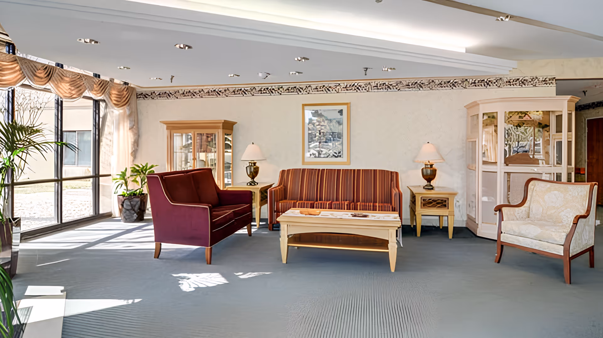 A bright and spacious living room area with large windows letting in natural light. The room features a striped sofa, a maroon loveseat, a cream-colored armchair, two wooden side tables with lamps, a wooden coffee table, and two glass display cabinets. There are plants near the windows and a framed picture on the wall.