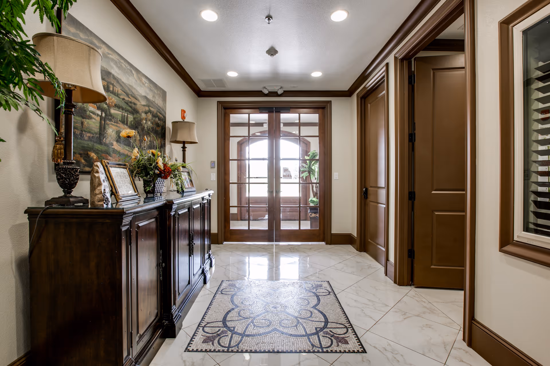 A well-lit interior hallway with polished white marble floors featuring a decorative tile pattern in the center. On the left side, there is a dark wooden cabinet adorned with two table lamps, framed pictures, and a floral arrangement. Above the cabinet hangs a large landscape painting. At the end of the hallway, there are double glass doors with wooden frames leading to another room with a large window and a potted plant. The walls and door frames are painted in neutral tones with brown trim.