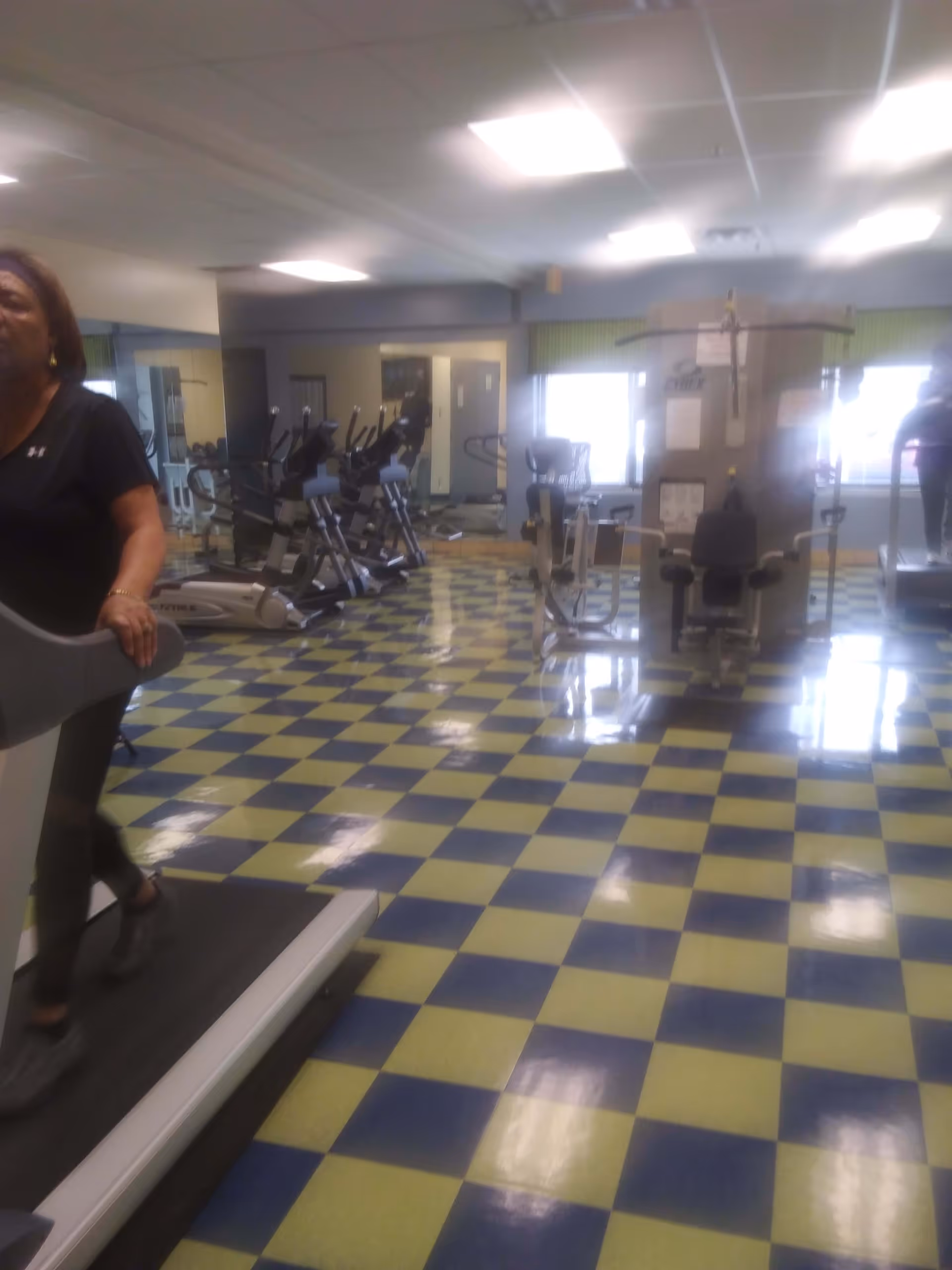 Indoor gym area with exercise equipment including treadmills, stationary bikes, and weight machines. A woman is walking on a treadmill on the left side of the image. The floor has a blue and yellow checkered pattern, and the room is well-lit with ceiling lights and windows in the background.