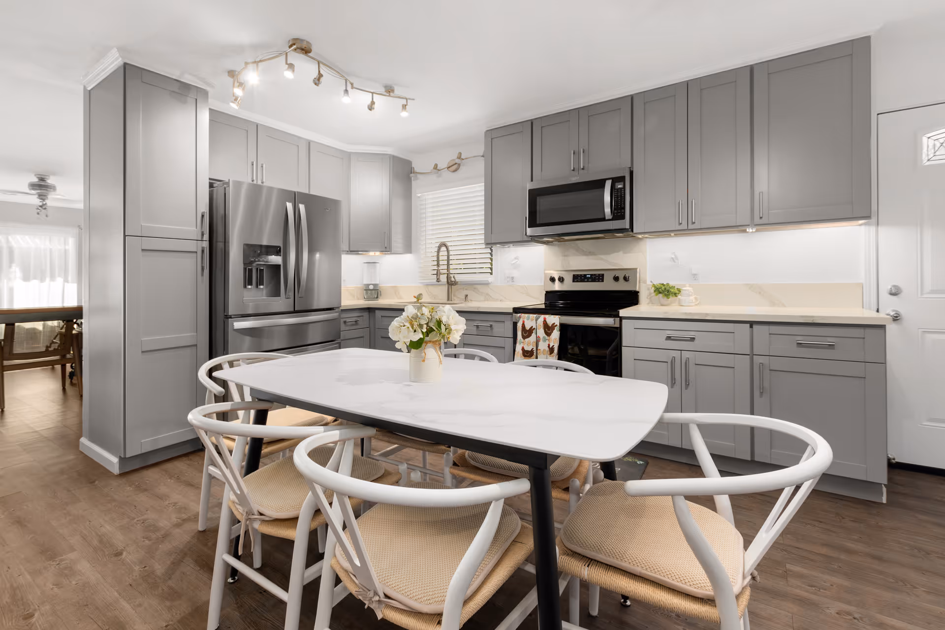 Modern kitchen with gray cabinets, stainless steel refrigerator, stove, and microwave. A white dining table with six chairs is in the center, and a vase with flowers is on the table. The floor is wooden, and there is a window with blinds above the sink.