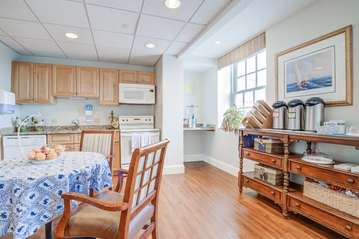 A bright kitchen and dining area featuring wooden cabinets, a white stove and microwave, a dishwasher, and a granite countertop. A table covered with a blue and white patterned tablecloth is surrounded by wooden chairs. On the right side, a wooden sideboard holds coffee dispensers, baskets, and other items. A large window lets in natural light, and a framed painting of a sailboat hangs on the wall above the sideboard.
