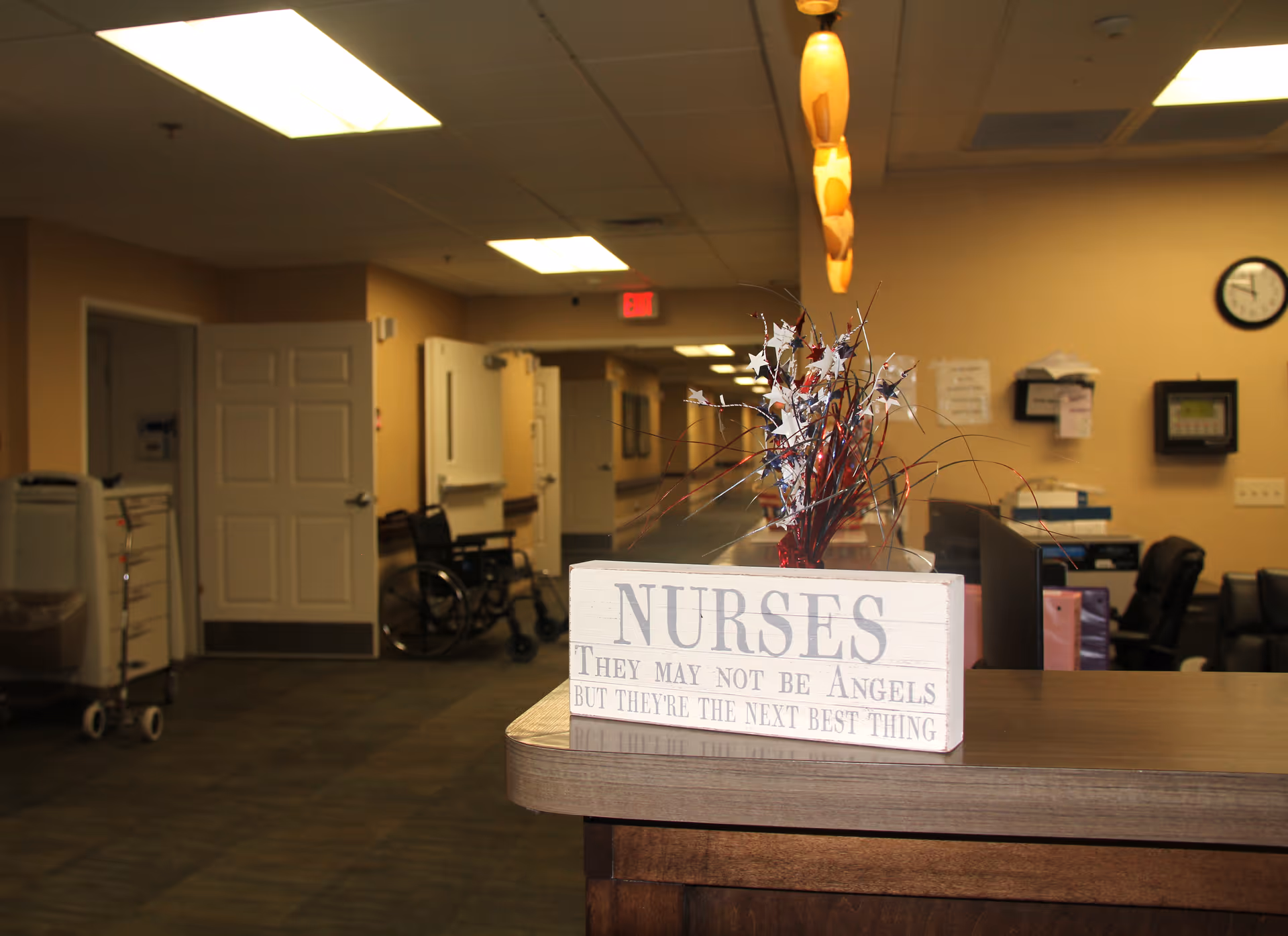 Reception desk area in a healthcare facility with a decorative sign that reads 'NURSES They may not be Angels but they're the next best thing.' In the background, there is a hallway with open doors, a wheelchair, and medical equipment. The walls are beige, and the ceiling has fluorescent lights.