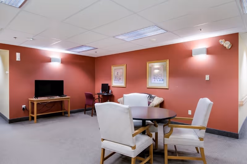 A common area with a round table surrounded by four cushioned chairs, a TV on a wooden stand against a red wall, a small desk with a computer and chair, and two framed pictures hanging on the wall.