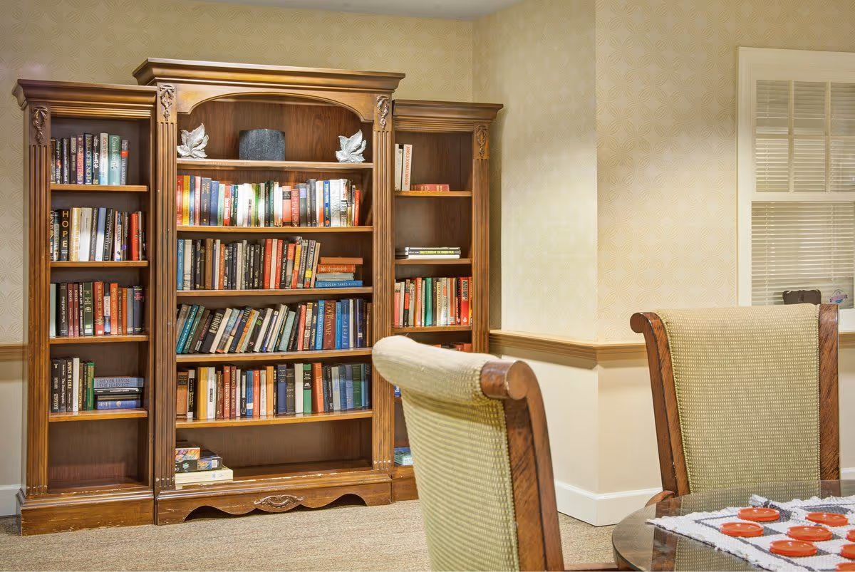 A cozy room corner featuring a large wooden bookshelf filled with books. In the foreground, there is a cushioned chair with wooden armrests and a glass table with a checkers game set on a crocheted tablecloth. The walls have light-colored patterned wallpaper and a window with white blinds.