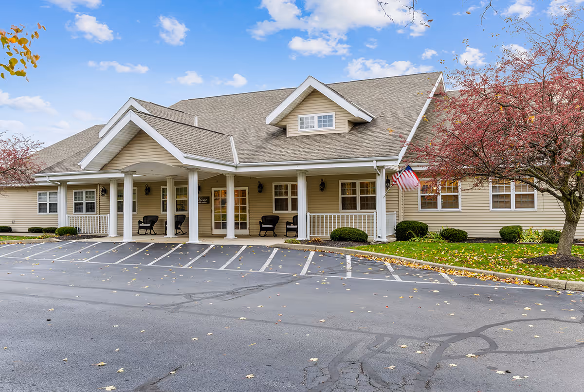 Front exterior of a single-story beige senior living building with a covered entrance, columns, seating, and an American flag.