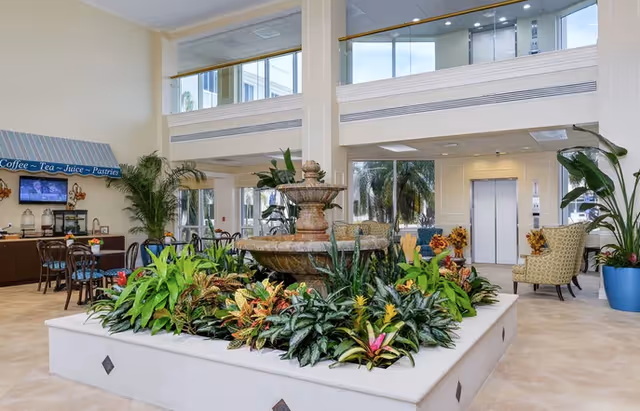Bright two-story interior lobby featuring a central planted fountain, seating areas, and a coffee station.