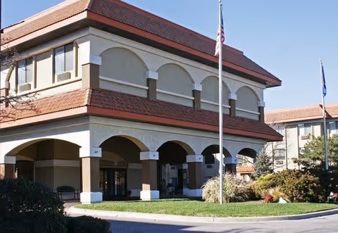 Exterior view of Sedgwick Plaza building showing a two-story structure with arched architectural details, a red-tiled roof, and two flagpoles with flags in front. The building is surrounded by landscaped greenery and a driveway.
