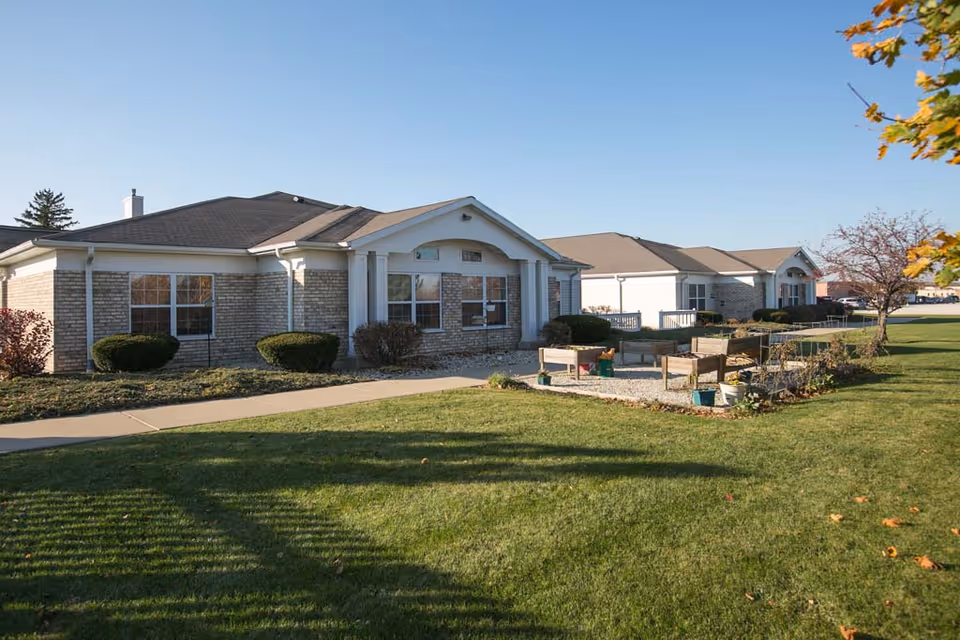 Exterior view of a single-story senior living facility building with brick and light-colored siding, surrounded by a well-maintained lawn and small garden beds under a clear blue sky.