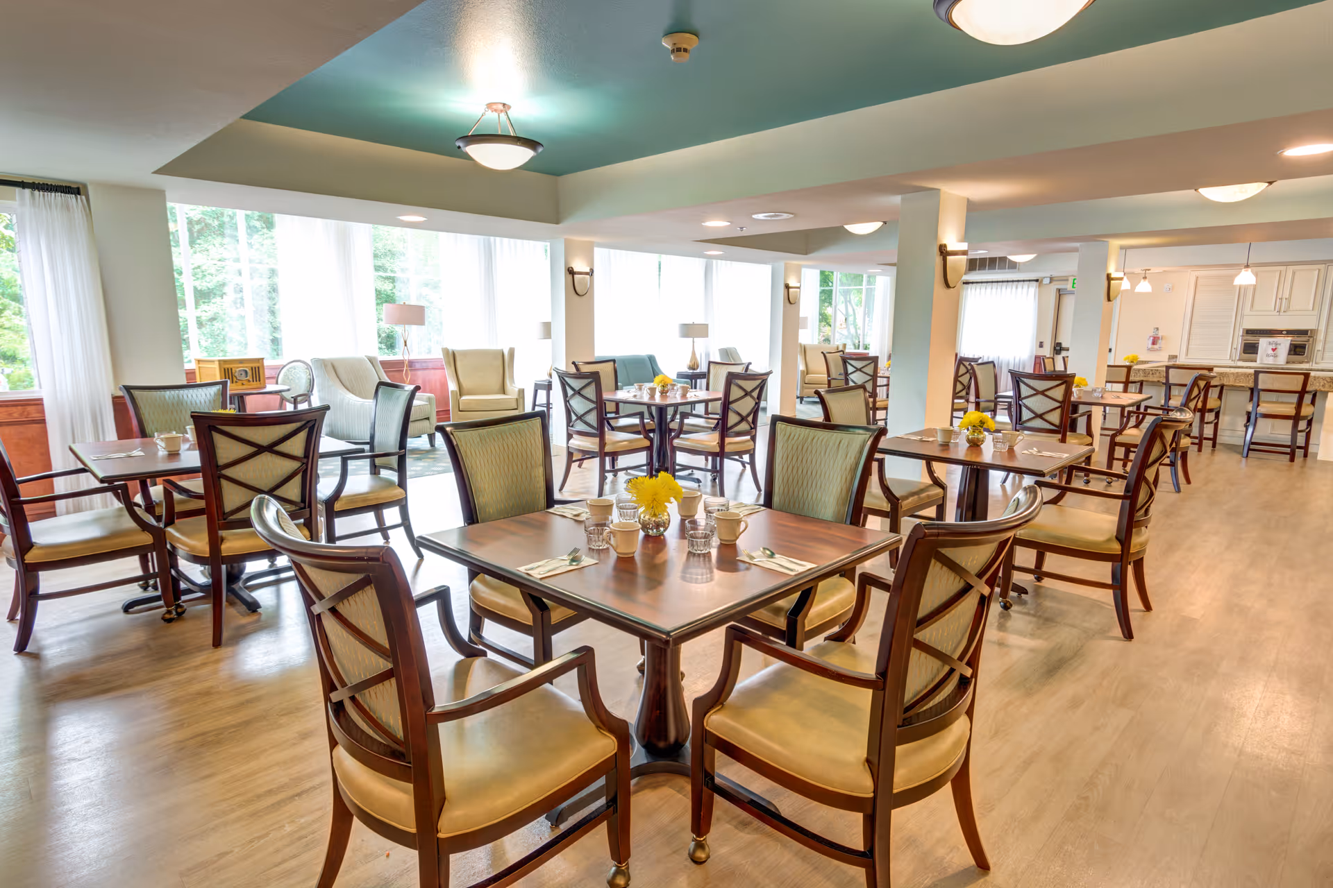A bright and spacious dining room in a senior living facility with multiple wooden tables and chairs arranged neatly. Each table has a small flower vase with yellow flowers and place settings. Large windows with sheer white curtains allow natural light to fill the room. The room has a light wood floor and a ceiling with recessed lighting and a central light fixture.