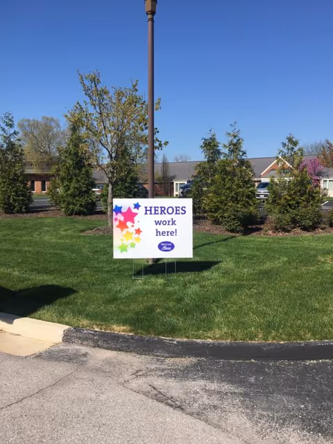 Outdoor view of a grassy area with a sign that reads 'HEROES work here!' in front of a lamp post, surrounded by trees and a building in the background under a clear blue sky.