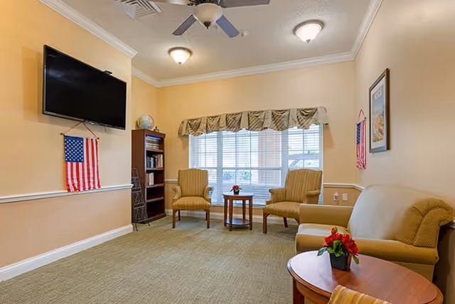A cozy living room area with beige walls and carpet, featuring a wall-mounted flat screen TV, two striped armchairs and a small wooden table with a flower vase in front of a large window with blinds and a valance. There is a wooden bookshelf with books and a globe, an American flag hanging on the wall, and a beige sofa with a round wooden side table holding a flower vase.