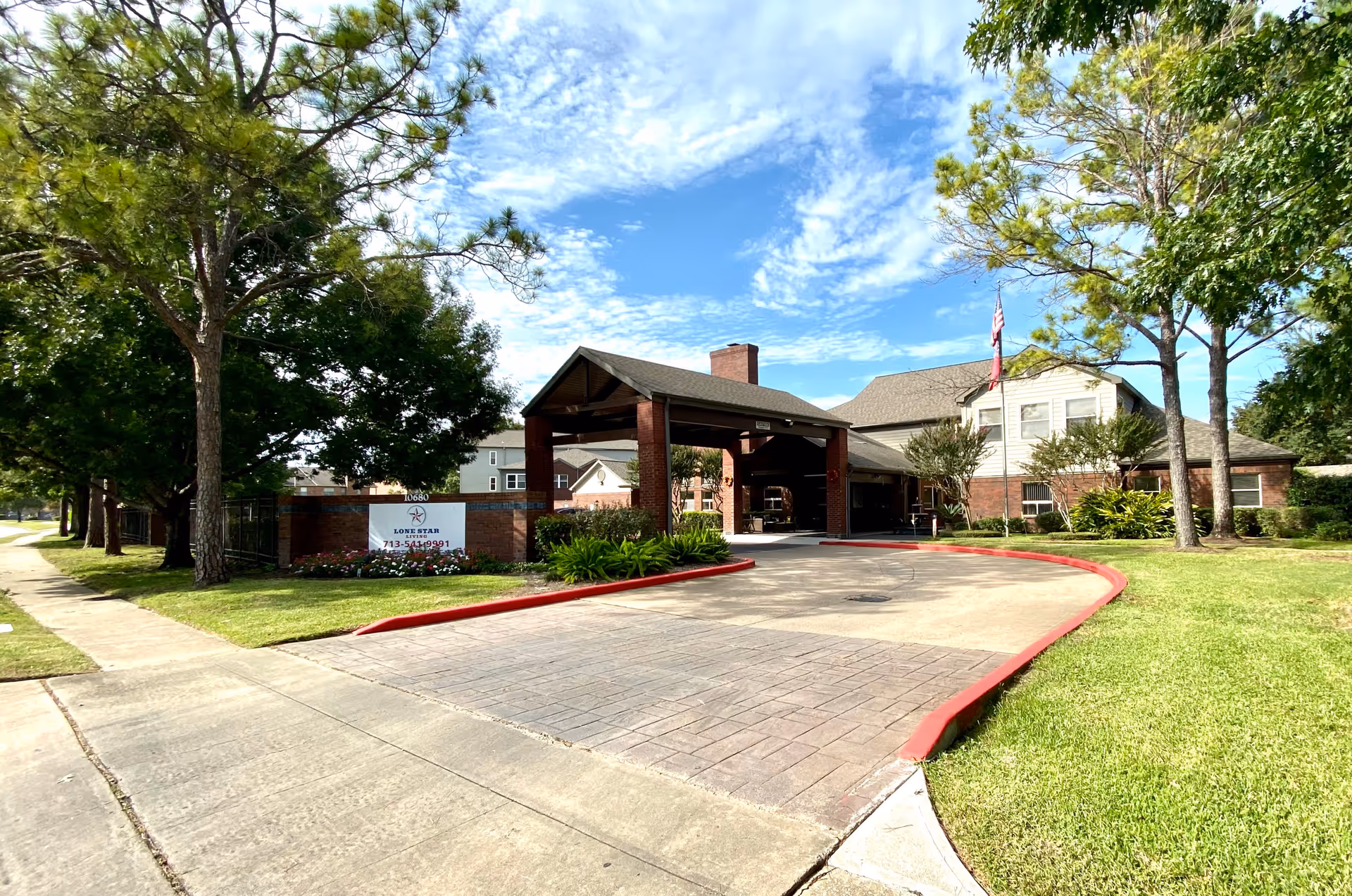 Front exterior view of Lone Star Living facility showing a covered entrance with brick pillars, a driveway with red curbs, green lawns, trees, and a clear blue sky with scattered clouds.