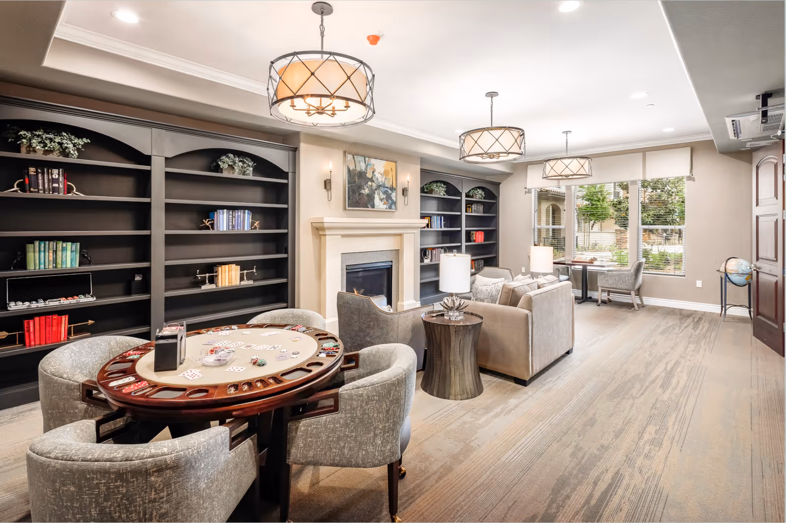 A bright and spacious senior living common area featuring a round game table with four upholstered chairs, a cozy seating area with a sofa and armchairs around a fireplace, large windows letting in natural light, built-in black bookshelves with decorative items, and modern ceiling lights.