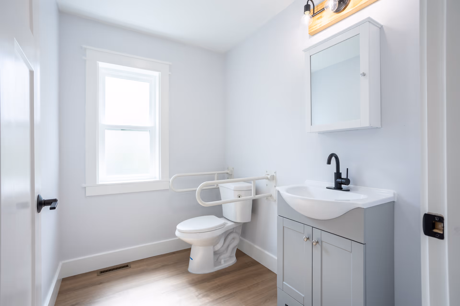 Bright accessible bathroom with a toilet and fold-down grab bars, a vanity sink with a mirror cabinet, and a window.