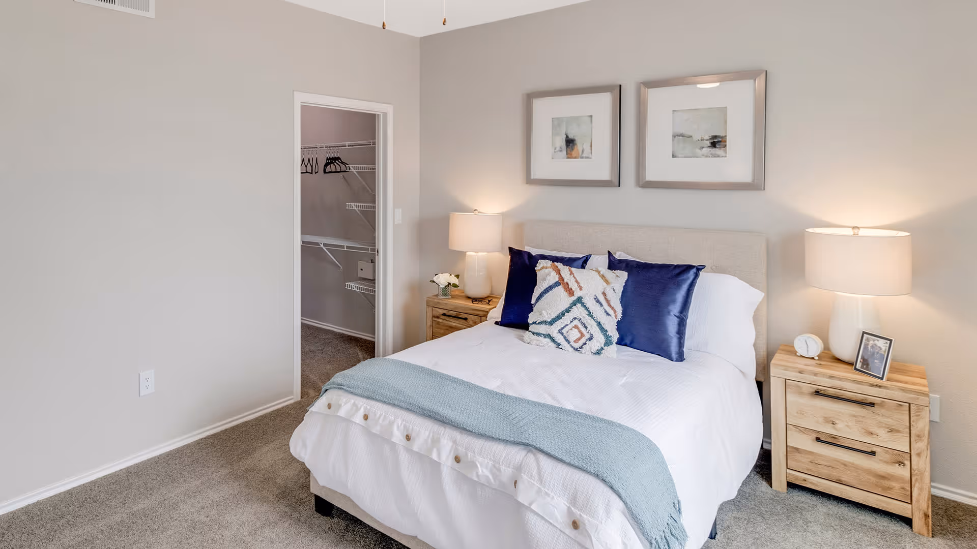 A bedroom with a neatly made bed featuring white bedding, a light blue throw blanket, and decorative pillows. There are two wooden nightstands on either side of the bed, each with a lamp. Two framed abstract artworks hang above the bed. An open doorway reveals a walk-in closet with shelves and hanging rods.