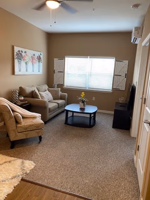 A cozy living room with a beige sofa and patterned armchair, a dark wooden coffee table with a small plant on it, a window with white blinds and decorative wooden shutters, a ceiling fan with light, a wall-mounted air conditioning unit, and a TV on a stand. The walls are painted light brown and there is a framed floral artwork above the sofa.