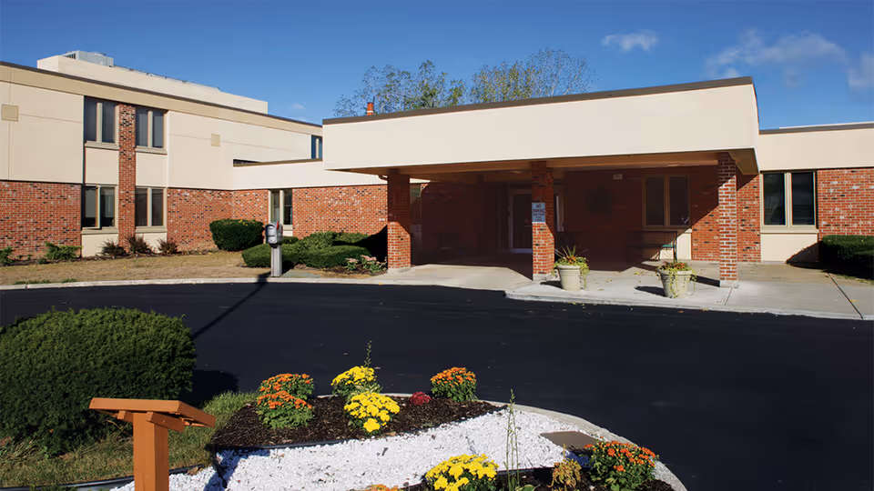 Front entrance of a brick and beige senior living building with a covered drive and landscaped flower bed.