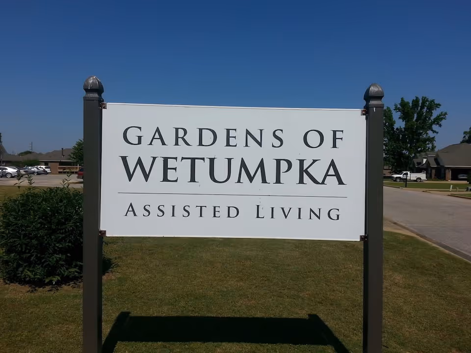 A white sign with black text that reads 'Gardens of Wetumpka Assisted Living' mounted on two dark posts, with a grassy area and a clear blue sky in the background.